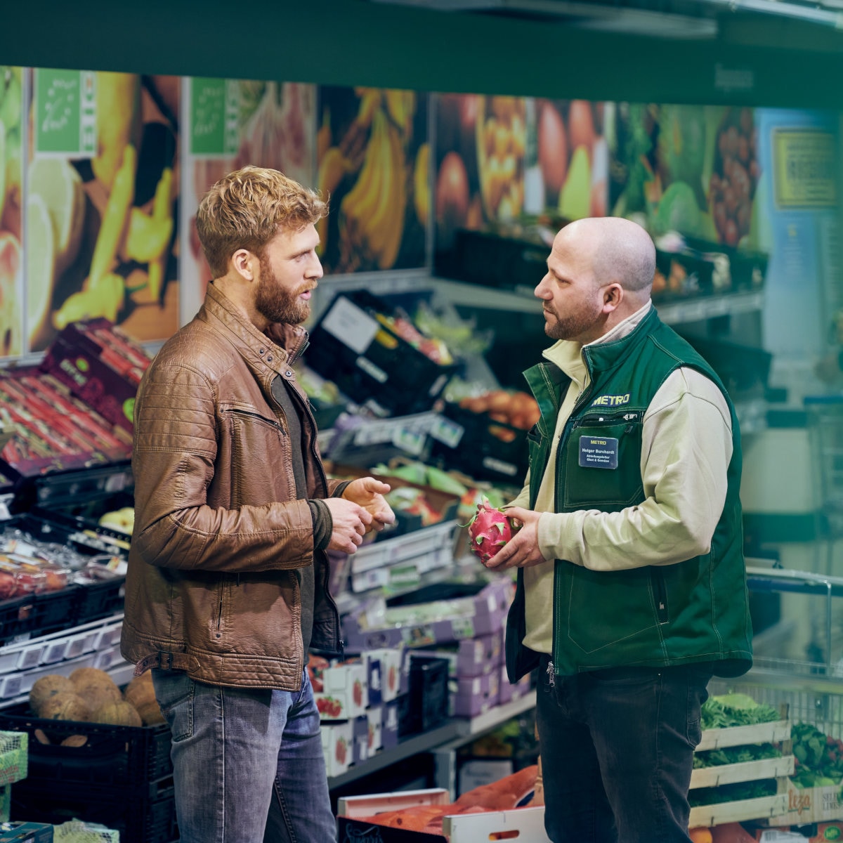 Two people discussing products in a grocery store produce section surrounded by fresh fruits and vegetables